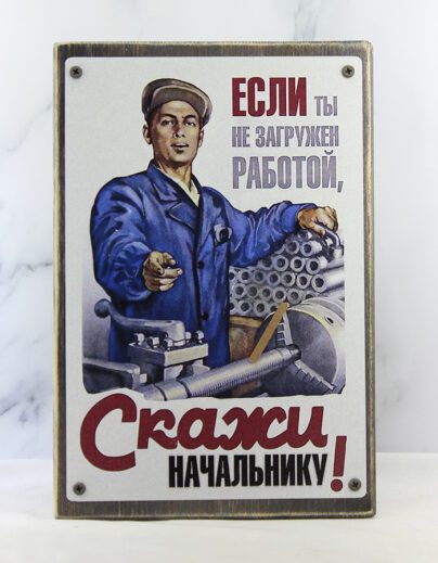 Рабочий в синем халате и кепке у токарного станка, рука на заготовках, указывает пальцем, надпись «Если ты не загружен работой, скажи начальнику» – демотиватор на состаренной доске